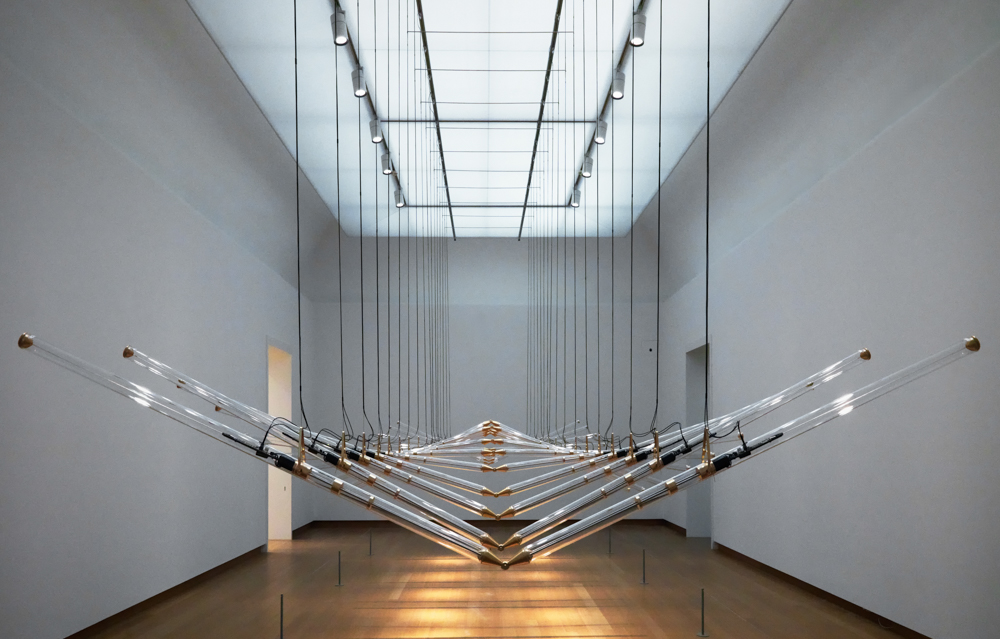 Undulating glass pipes forming repetitive geometric figures. The pipes hang from the ceiling in a large spacious room.