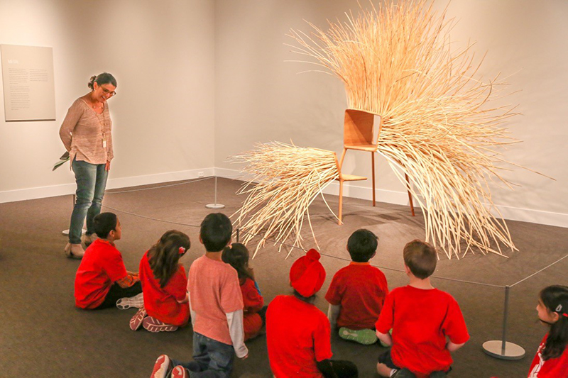 A group of children sit in front of a large wooden chair sculpture and listen to a woman who is standing up and teaching them about it
