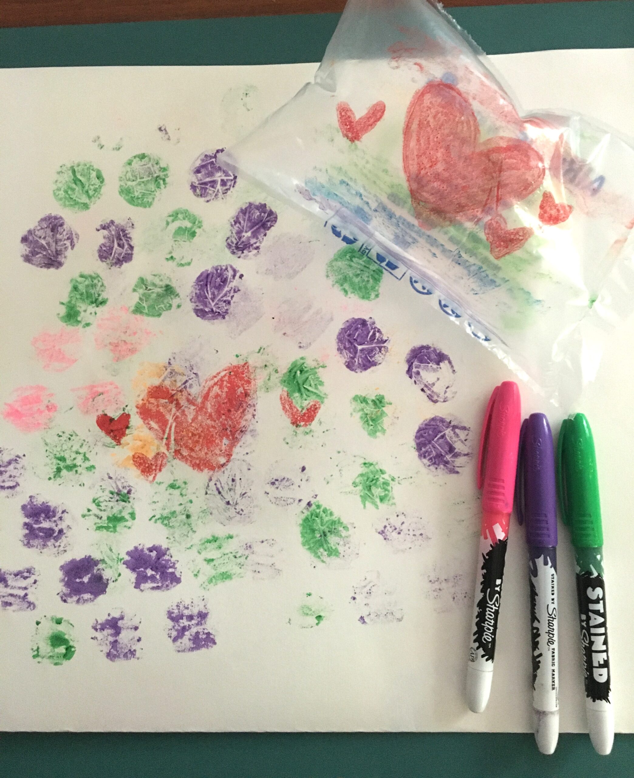 Markers and a plastic bag sitting next to a piece of paper decorated with dots and hearts transferred from the plastic bag.
