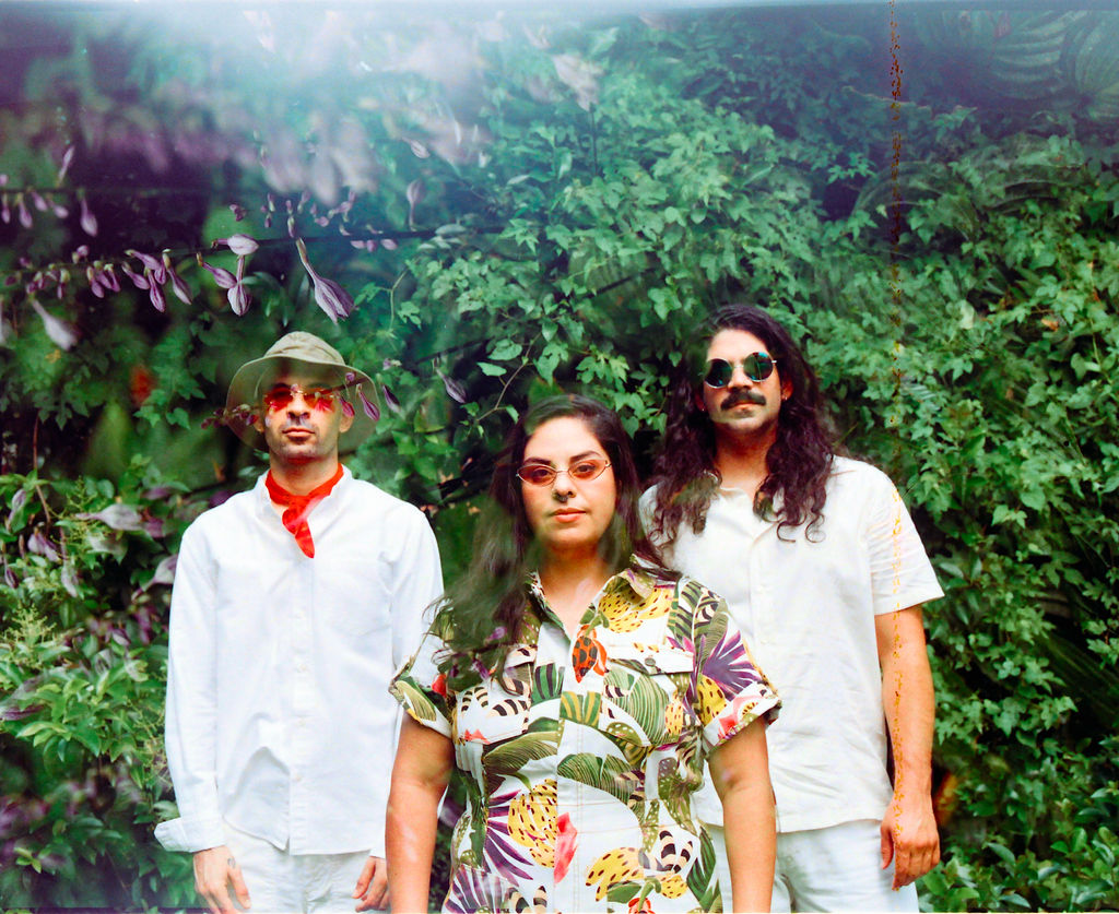 Two men standing dressed in white, one man wearing a hat and red bandana. One woman standing between the two mean dressed in a jungle-print shirt. All are standing in front of a lush green plant background.