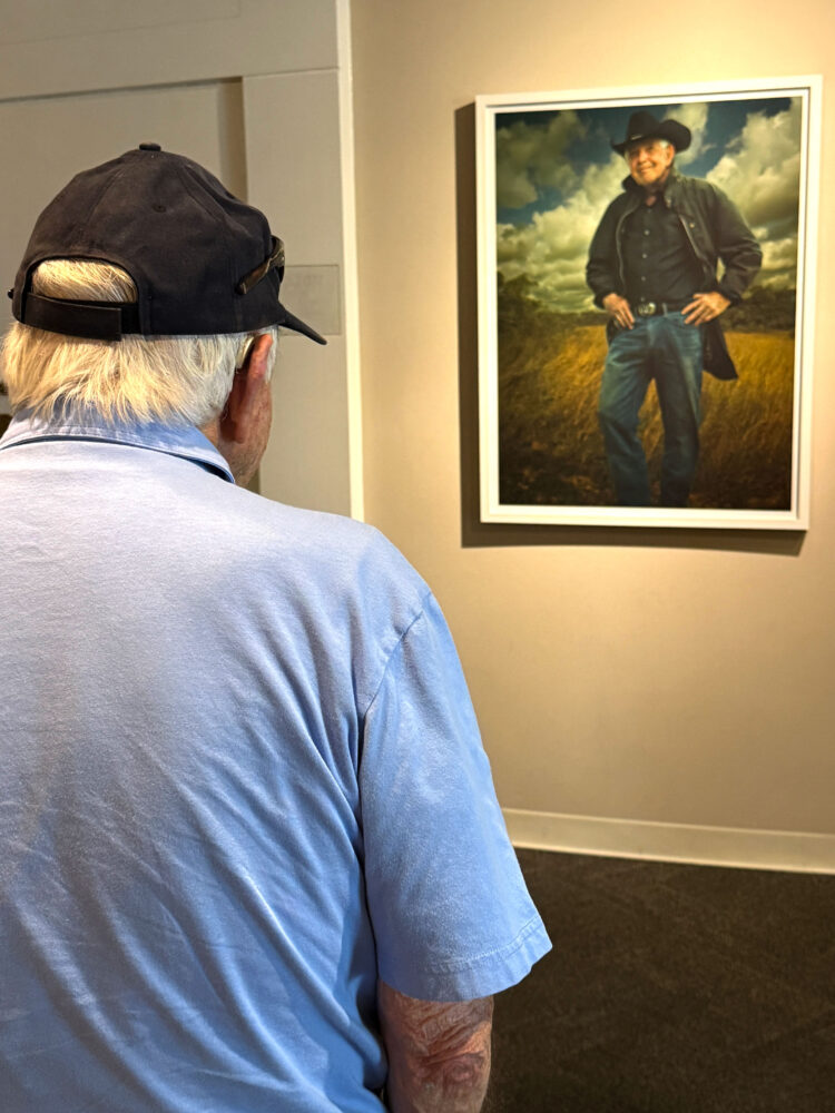 Man in blue short-sleeve shirt with his back to camera looks at portait of himself hanging on gallery wall.
