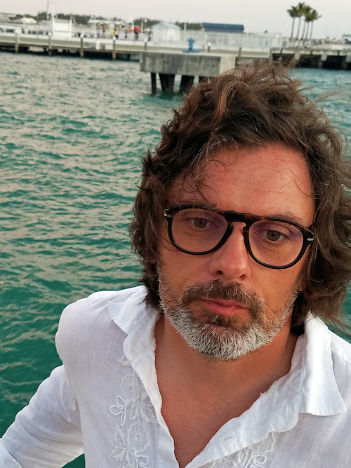 Man with dark hair wearing glasses by waterfront with wind blowing hair.